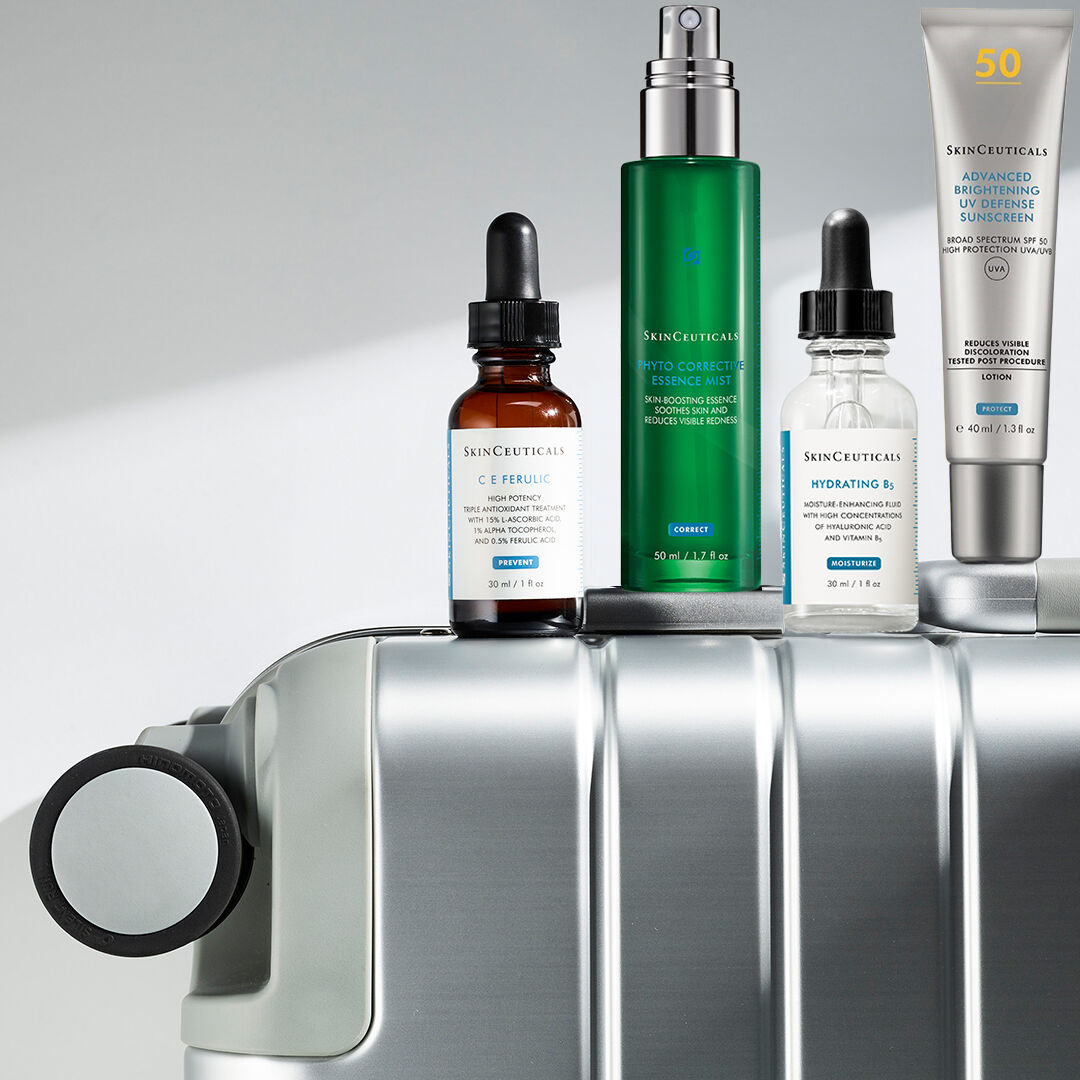 A selection of SkinCeuticals skincare products displayed on a silver suitcase, including a vitamin C serum, a hydrating serum, a corrective essence mist, and a sunscreen.