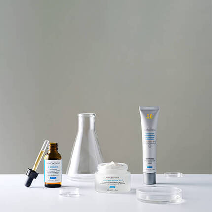 A clean, minimalistic arrangement of skincare products including a serum in a dropper bottle, a cream in a jar, and a sunscreen in a tube, placed alongside laboratory glassware on a white surface.