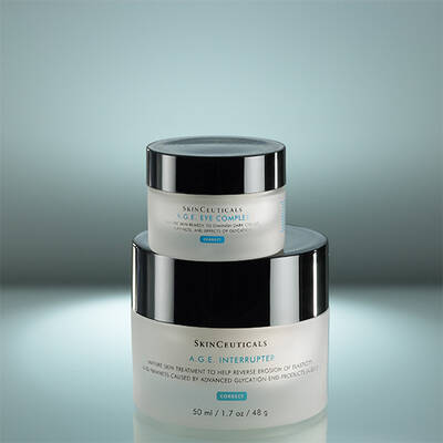 Two jars of SkinCeuticals skincare products are stacked on a reflective surface. The top jar is labeled "A.G.E. Eye Complex" and the bottom jar is labeled "A.G.E. Interrupter." The background is softly lit, emphasizing the jars' elegant design.