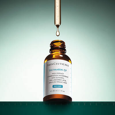 A dropper releasing a drop of liquid from a brown glass bottle of SkinCeuticals Silymarin CF, set against a gradient turquoise background.