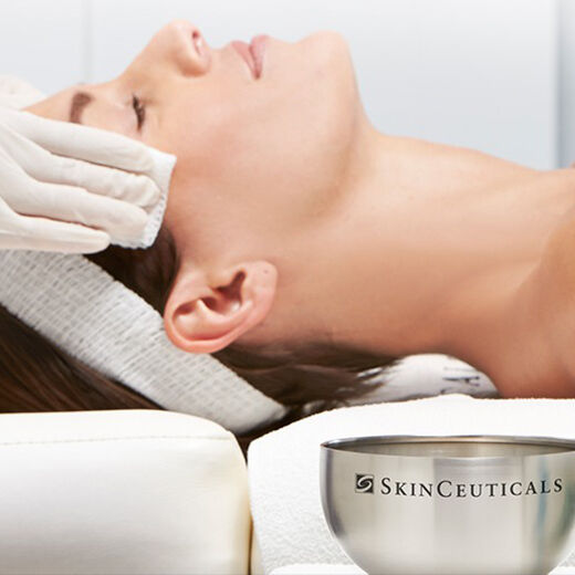 A woman receiving a facial treatment, lying on a table with her eyes closed, while a person wearing gloves gently applies a cloth to her forehead.