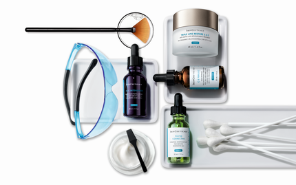 An assortment of skincare products, including serums and a moisturizer, arranged on a tray. Tools such as a fan brush and cotton swabs are also present. The SkinCeuticals logo is displayed in the top left corner.