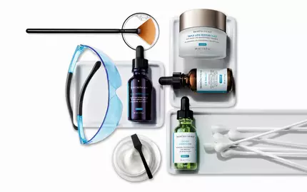 An assortment of skincare products, including serums and a moisturizer, arranged on a tray. Tools such as a fan brush and cotton swabs are also present. The SkinCeuticals logo is displayed in the top left corner.