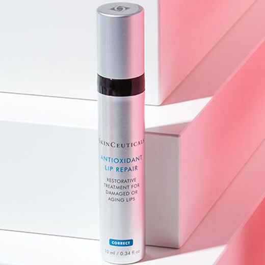 A silver tube of Antioxidant Lip Repair from a skincare brand, positioned against a backdrop of white and pink geometric shapes.