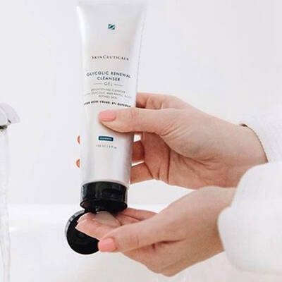 A person is dispensing a gel cleanser from a tube into their hand while standing near a bathroom faucet with running water.