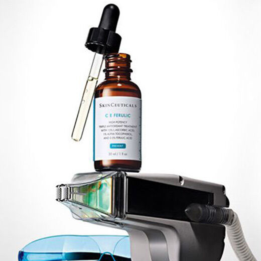 A dropper bottle of SkinCeuticals C E Ferulic serum is perched on top of a sleek skincare device. The bottle is brown glass with a white label, and a clear dropper extends from the top. The background is minimalist and neutral.