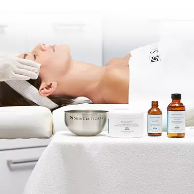 A woman receiving a facial treatment in a spa setting, with skincare products from SkinCeuticals displayed in the foreground. A technician is gently applying a product to the woman's face.