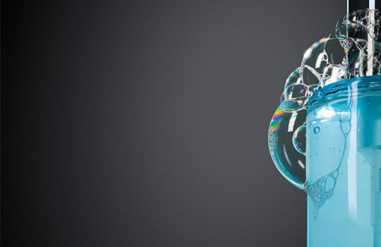 A close-up of a blue bottle with a pump, featuring bubbles forming around the nozzle against a dark backdrop.