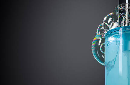 A close-up of a blue bottle with a pump, featuring bubbles forming around the nozzle against a dark backdrop.