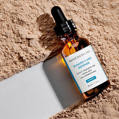 A close-up of a bottle of SkinCeuticals Blemish + Age Defense, a skin treatment product, resting on a textured surface with a neutral background.