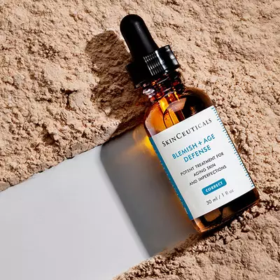 A close-up of a bottle of SkinCeuticals Blemish + Age Defense, a skin treatment product, resting on a textured surface with a neutral background.