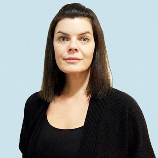 A woman with straight dark hair and light skin stands in front of a light blue background, wearing a black top and cardigan. She has a neutral expression.