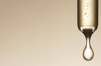Three glass droppers, each containing a clear liquid, hang against a soft beige background, dripping at varying intervals.