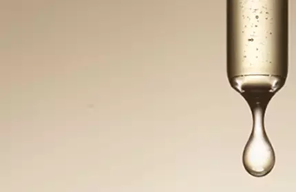 Three glass droppers, each containing a clear liquid, hang against a soft beige background, dripping at varying intervals.
