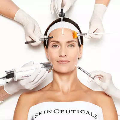 A woman is receiving a facial treatment, with various beauty tools being held around her face by gloved hands. She has a calm expression, and her hair is pulled back with a headband.