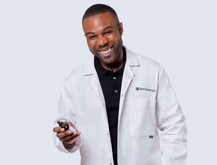 A smiling man wearing a white lab coat holds a small device, standing against a light blue and black background.