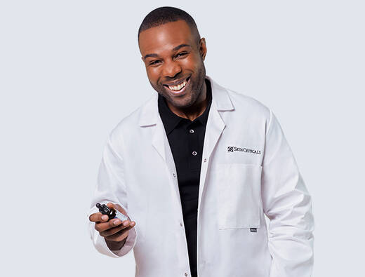 A smiling man wearing a white lab coat holds a small device, standing against a light blue and black background.