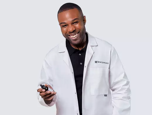 A smiling man wearing a white lab coat holds a small device, standing against a light blue and black background.