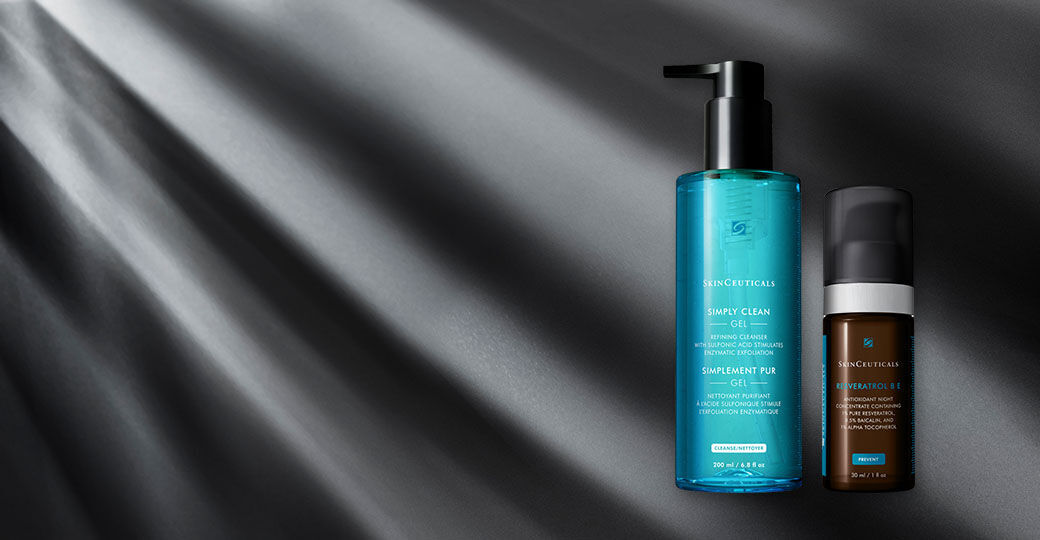 A bottle of SkinCeuticals Simply Clean Gel and a bottle of SkinCeuticals Retinol 0.5 sit on a dark surface with soft light rays shining down.