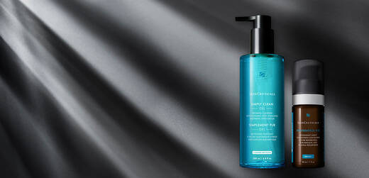 A bottle of SkinCeuticals Simply Clean Gel and a bottle of SkinCeuticals Retinol 0.5 sit on a dark surface with soft light rays shining down.