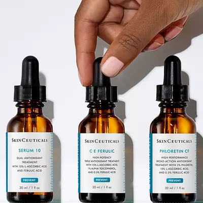 A hand reaches for a dropper bottle from a set of three skincare serums, all displayed against a white background. The bottles are labeled "Serum 10," "C E Ferulic," and "Phloretin CF."