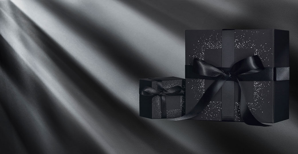 Two elegantly wrapped black gift boxes with shiny black ribbons sit against a dark background, illuminated by soft beams of light.