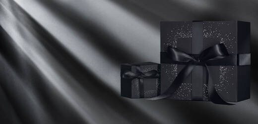 Two elegantly wrapped black gift boxes with shiny black ribbons sit against a dark background, illuminated by soft beams of light.