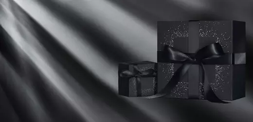 Two elegantly wrapped black gift boxes with shiny black ribbons sit against a dark background, illuminated by soft beams of light.