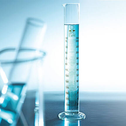 A clear graduated cylinder filled with blue liquid, positioned against a light background, with additional glassware partially visible in the foreground.
