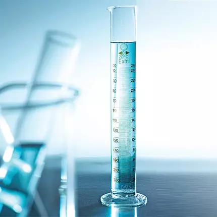 A clear graduated cylinder filled with blue liquid, positioned against a light background, with additional glassware partially visible in the foreground.