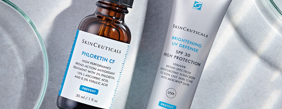 A close-up of two SkinCeuticals products: a brown glass bottle labeled "Phloretin CF" and a tube labeled "Brightening UV Defense SPF 30." The products are placed on a light surface, surrounded by small glass dishes.