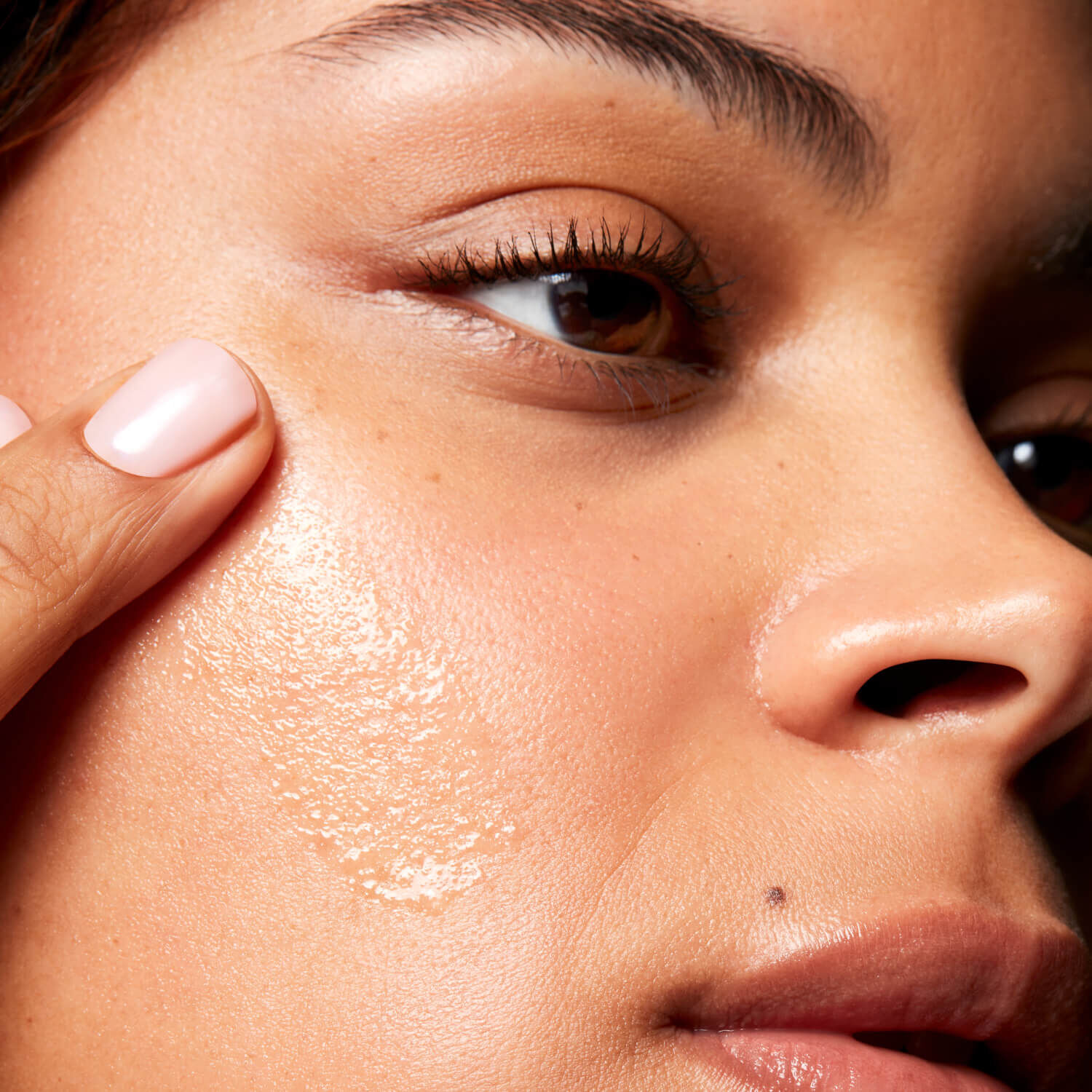 close up of a woman applying a pale gold serum to her cheek, looking into the distance