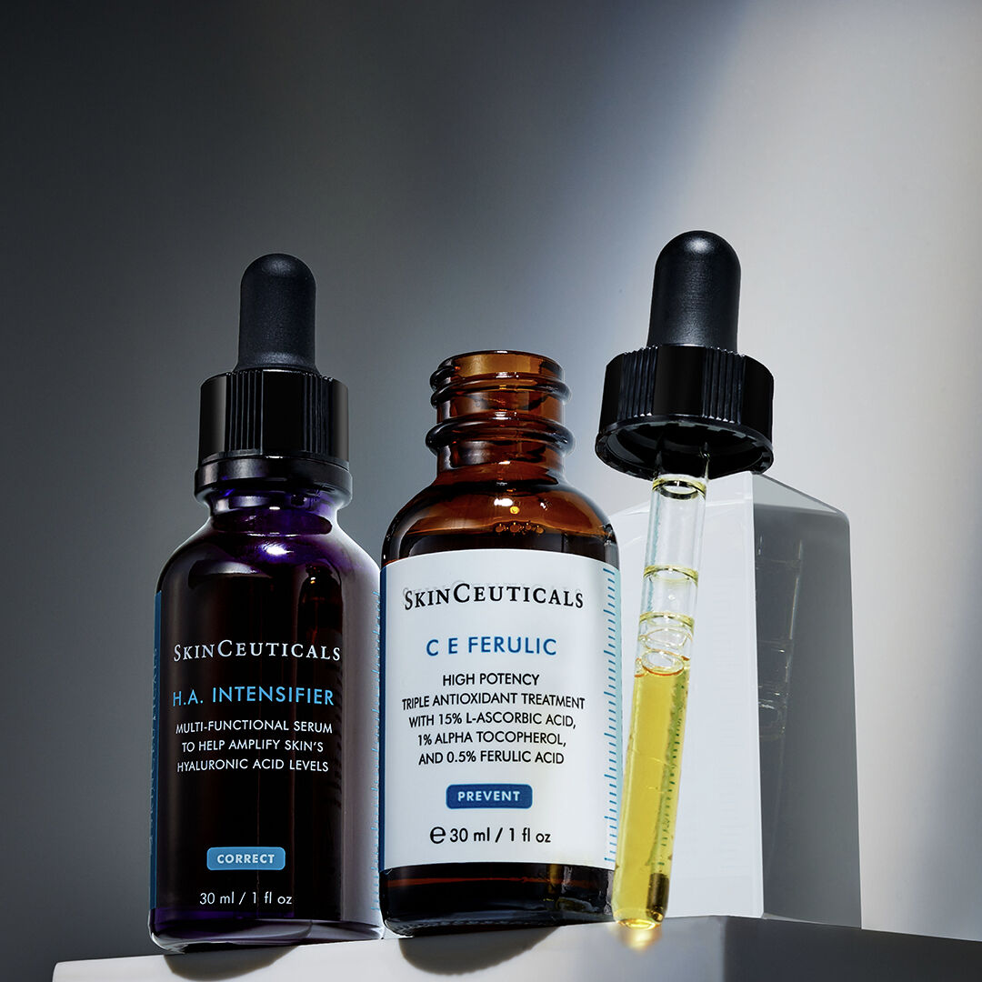 A bottle of C E Ferulic serum is open, its pipette lid resting beside it. Next to it sits a bottle of H.A. Intensifier serum in the shadows.