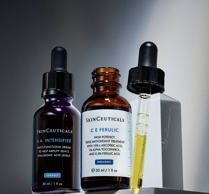 A bottle of C E Ferulic serum is open, its pipette lid resting beside it. Next to it sits a bottle of H.A. Intensifier serum in the shadows.