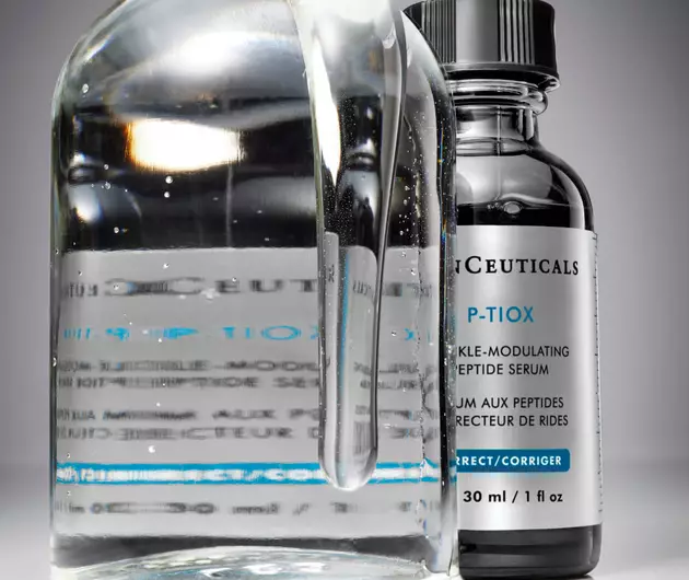 A glass bottle filled with clear liquid beside a dark serum bottle, both labelled with skincare product details.