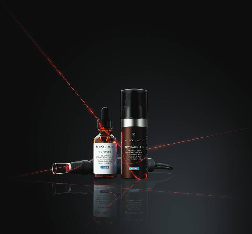 Two SkinCeuticals serums stand against a dark background. On the left, C E Ferulic serum, in an amber-coloured glass dropper bottle. On the right, Resveratrol B E serum, in a cylindrical brown pump bottle. A skincare tool rests next to them. Red lasers slice the darkness, adding a futuristic, high-tech edge.