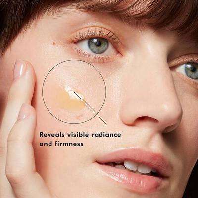 Close up: A pump of pale yellow serum sits untouched on a woman's cheek. Her hand rests on the side of her face.