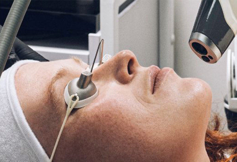 A woman lies on a treatment bed with an eye protection device, undergoing a skincare procedure in a clinical setting.