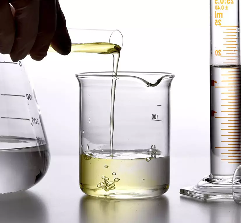 A hand pouring a yellow liquid from a small tube into a beaker, with measuring equipment in the background.